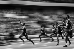 Black-and-white panned photo of male sprinters on a track with the leading runner airborne and raising his arm as competitors trail behind against a blurred stadium crowd.