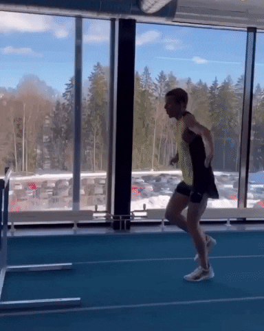 An athlete wearing a sleeveless top and shorts sprints along an indoor track and performs repeated running jumps over low hurdles beside floor-to-ceiling windows with snow and trees visible outside.