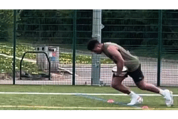 Athlete in a tank top and shorts explosively sprinting from a crouched start on an outdoor turf field with small orange cones marking the drill and a metal fence in the background.