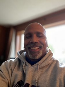 Smiling bald man with salt-and-pepper beard wearing a gray hoodie and earbuds, seated indoors by a bright window.