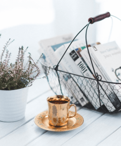 Golden teacup and saucer with tea on a white wooden table, small potted plant and wire basket of magazines behind.